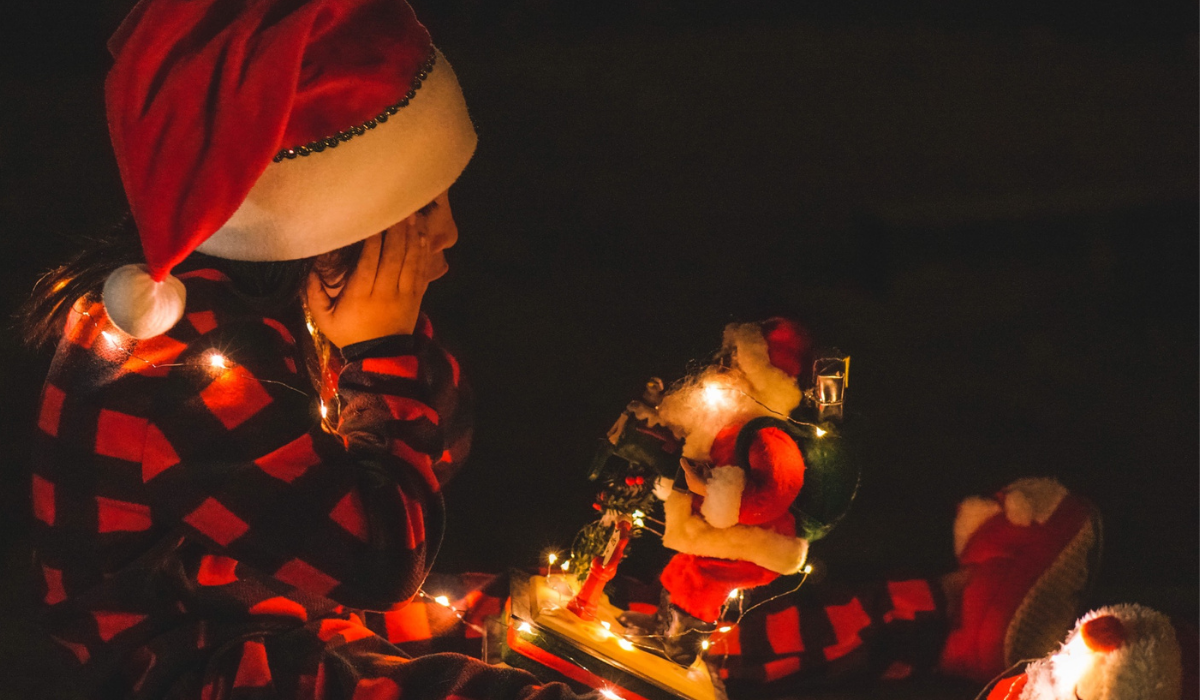 A young boy sitting in the dark with his legs out in front of him. He has a Santa Claus figure in his lap and holding his cheeks with his hands in disbelief at the lit up figurine.